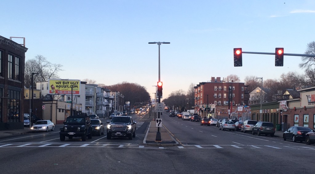 A wide multi-lane street with cars waiting at a red traffic light dusk.