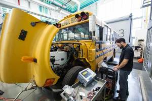 A technician works on a laptop next to an electric yellow school bus with its front hood opened for maintenance.