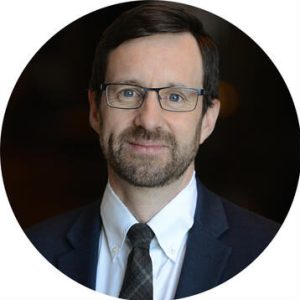 A headshot of a middle-aged white man with closely cropped facial hair and rectangular glasses wearing a suit and tie. 