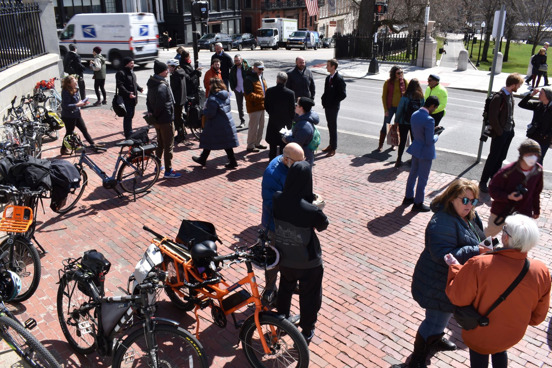 A crowd gathers in front of the Massachusetts State House next to a handful of pedal-assist electric bikes. 