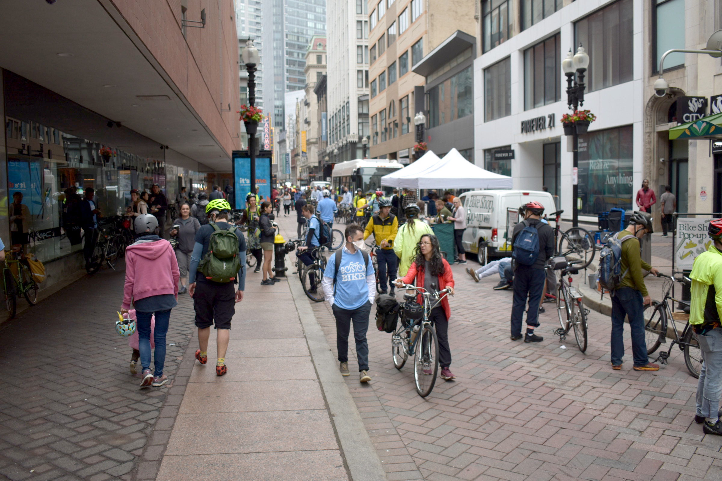 A crowd of Bike to Work Day celebrants, many of whom are wearing bike helmets, gather in front of tents to get free coffee and pastries in Downtown Crossing. 
