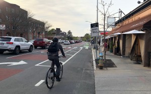 A bike rider rides along a flexpost-protected bike lane next to a sidewalk cafe on one side and a line of cars waiting in traffic on the other side of the bus lane, on the left edge of the photo.
