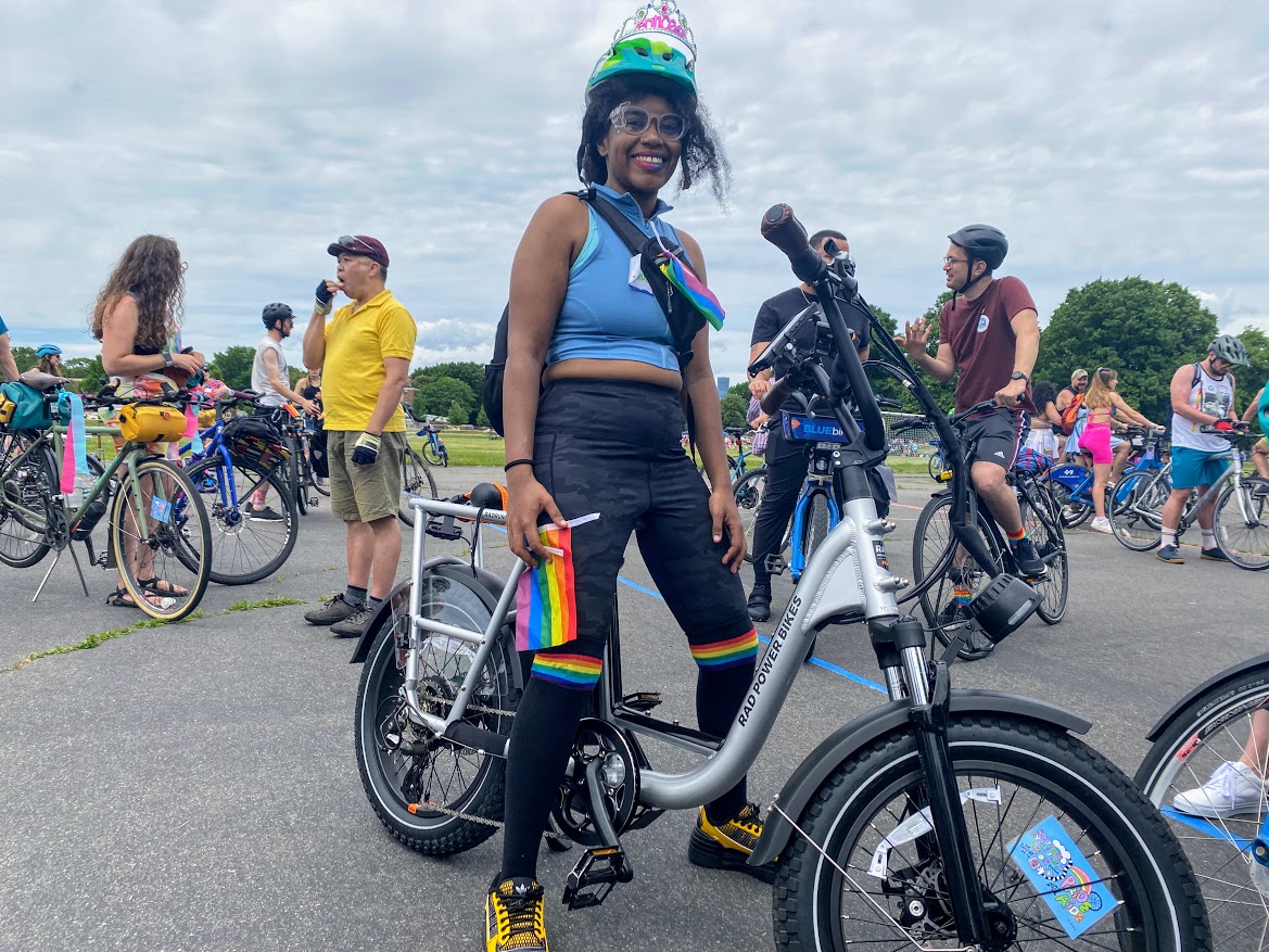 person wearing a blue shirt and black pants with colorful pride bands posing on her electric bike