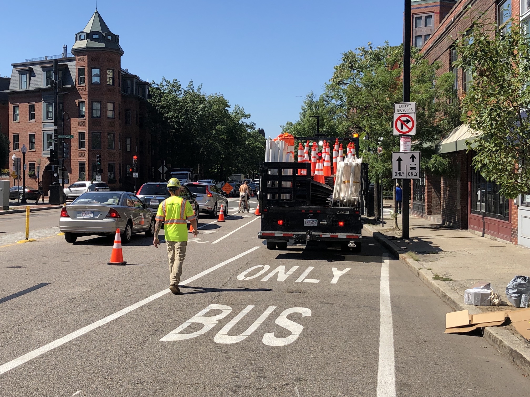 A truck full of construction cones is parked in a lane marked "BUS ONLY" while a worker in a yellow vest sets out cones to delineate the lane from general motor vehicle traffic