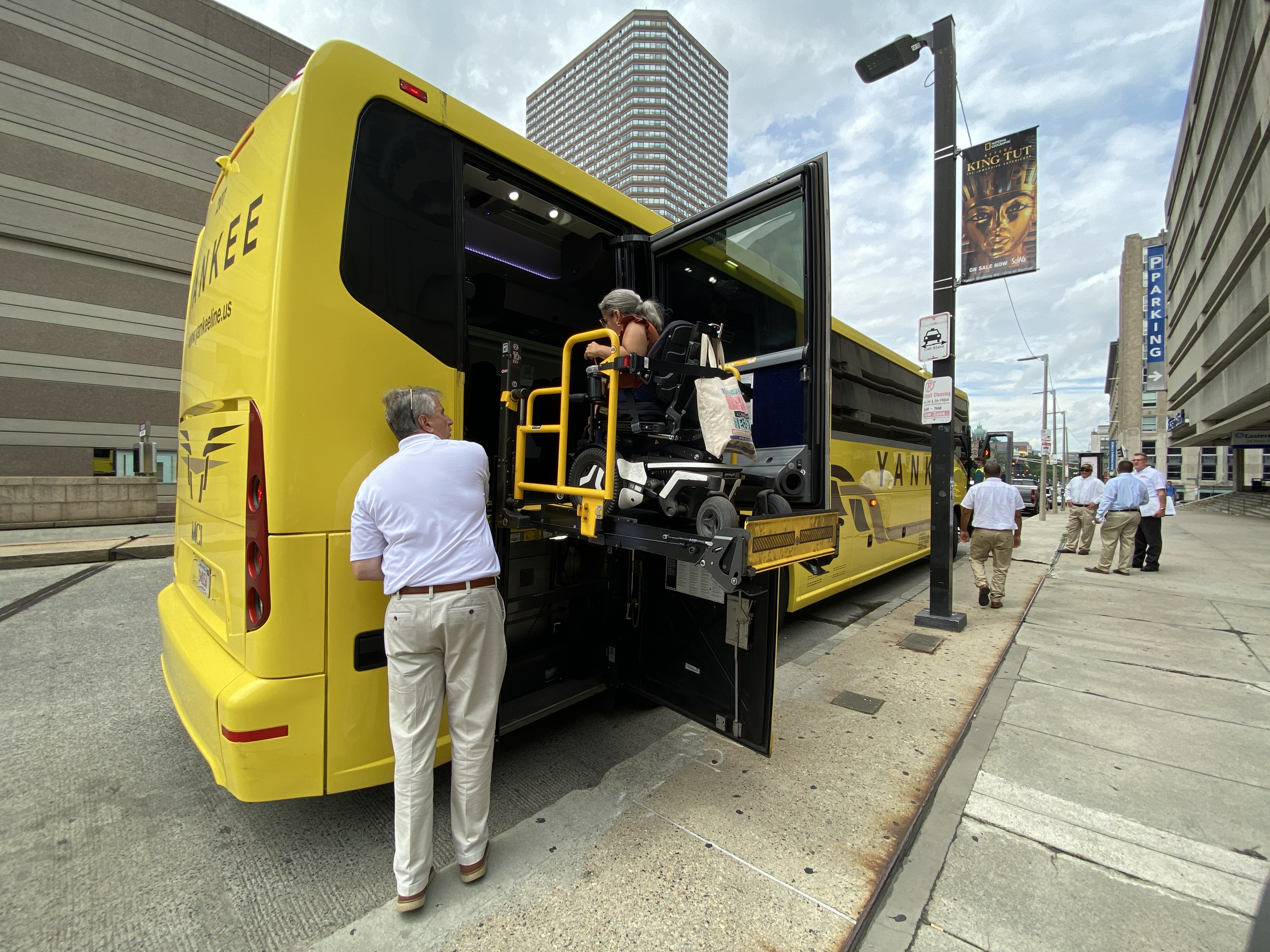 A wheelchair user rides on a lift to board a yellow Yankee Line bus from its rear door while an attendant assists from the sidewalk.