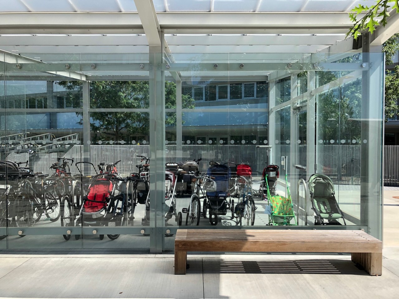 About a dozen baby strollers stored inside a glass-clad bike parking shed next to bike racks.