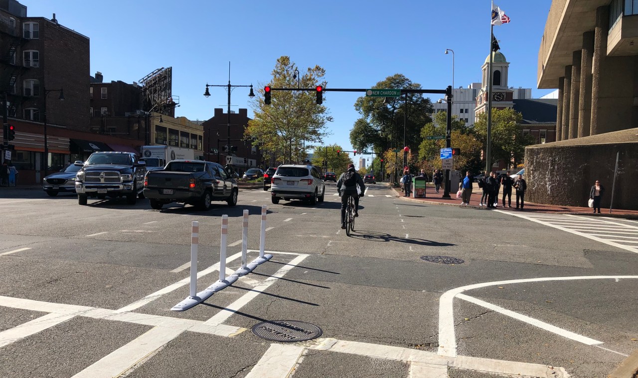 A bicycle rider coasts through an intersection in a bike lane next to two cars and a pickup truck waiting to turn left from the oncoming direction. In the foreground, a row of flexible-post bollards separates the bike lane from the car lanes. A group of pedestrians on the opposite corner waits to cross the street.