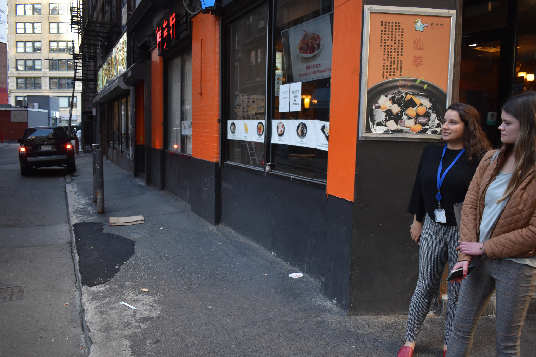 Two women stand on a bumpy asphalt sidewalk pockmarked with asphalt patches.