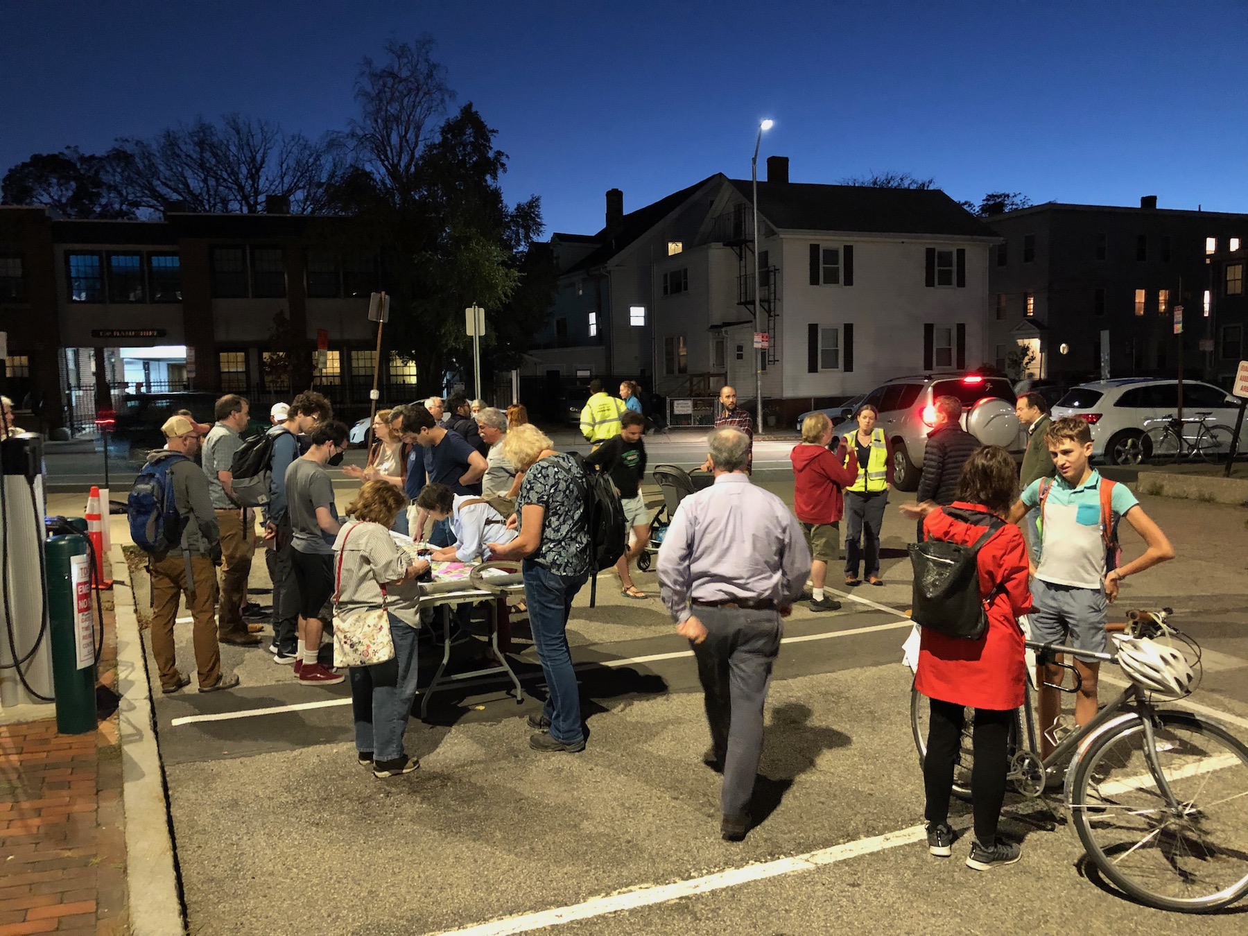A crowd of people gathered in a parking lot to discuss plans for Hampshire Street. Most of the crowd is gathered around a table on the left side of the photo, where a large map of the street is on display. Other people talk with each other in small groups in other parts of the parking lot. Several attendees are carrying bikes or strollers.