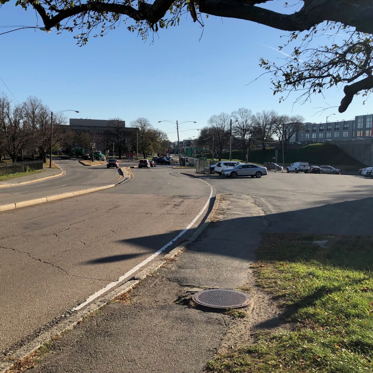 A broken, potholed asphalt sidewalk runs between a parking lot and a wide, mostly empty 4-lane roadway, both areas with much smoother pavement. 