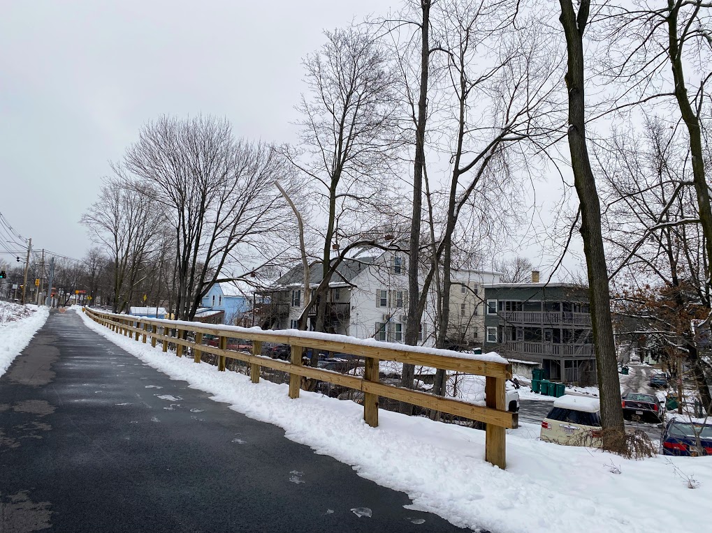 paved trail with some patches of slush and snow along either side. a wooden fence separates it from neighborhood homes.