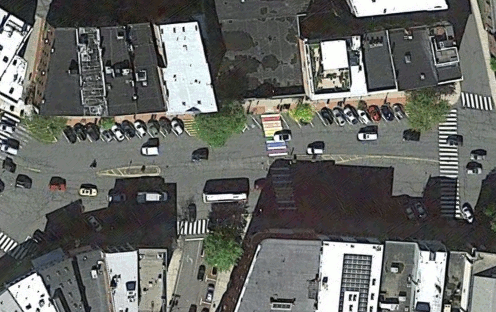 An animated GIF shows an overhead view of Northampton's Main Street between Crafts Avenue and Center Street. One frame of the animation shows the current satellite view, and the next shows the same view overlaid by MassDOT's design drawings for an upcoming construction project that will widen sidewalks, reconfigure on-street parking, and add protected bike lanes with a narrowed roadway for motor vehicles. 