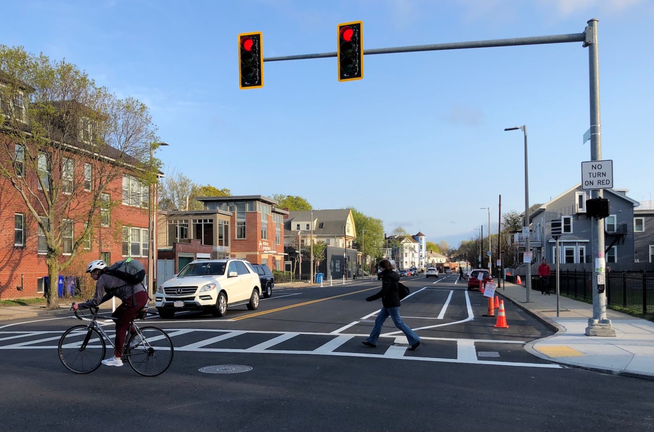 A view down a city street. On the right, cones block people from parking in the work zone where city workers are installing new lane striping and flexible-post bollards to designate a new protected bike lane.