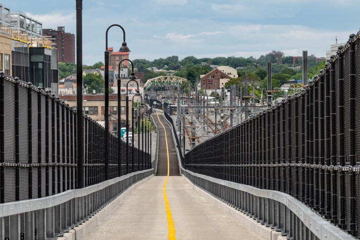 A view down a straight pathway with a yellow line painted down its center. The path is lined with black fencing and railroad infrastructure is visible to the right. In the distance is a bridge over the tracks and an MBTA station sign, and a skyline of buildings and trees
