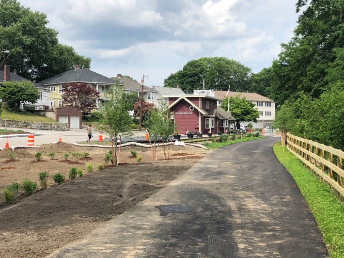 A freshly-paved trail through a suburban neighborhood. Newly-planted trees line the edge of the path, and to the right is a new wooden fence. In the middle distance is an old train station with the sign "Waltham Highlands"