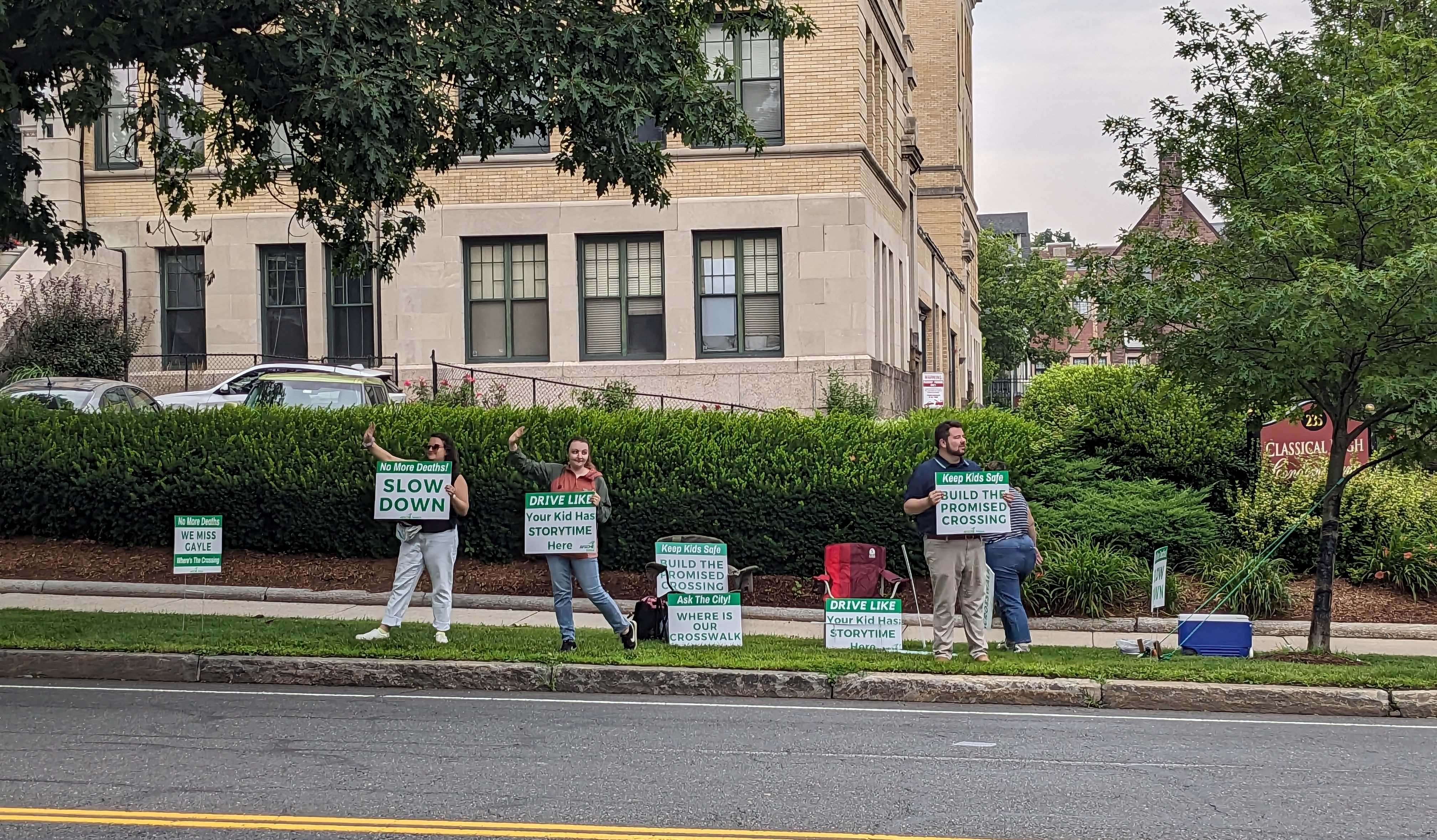 Four people hold green-and-white signs along the curb of a wide four-lane roadway. A grand Italianate-style library building with tall arched windows is in the background behind them on the other side of the street. The man on the right holds a sign reading "keep kids safe: build the promised crossing." One woman to the left holds a sign that reads "Drive like your kid has storytime here" and the woman at the left edge of the photo holds a sign reading "No more deaths! SLOW DOWN." A fourth person on the right is walking towards the sidewalk behind the people holding signs.