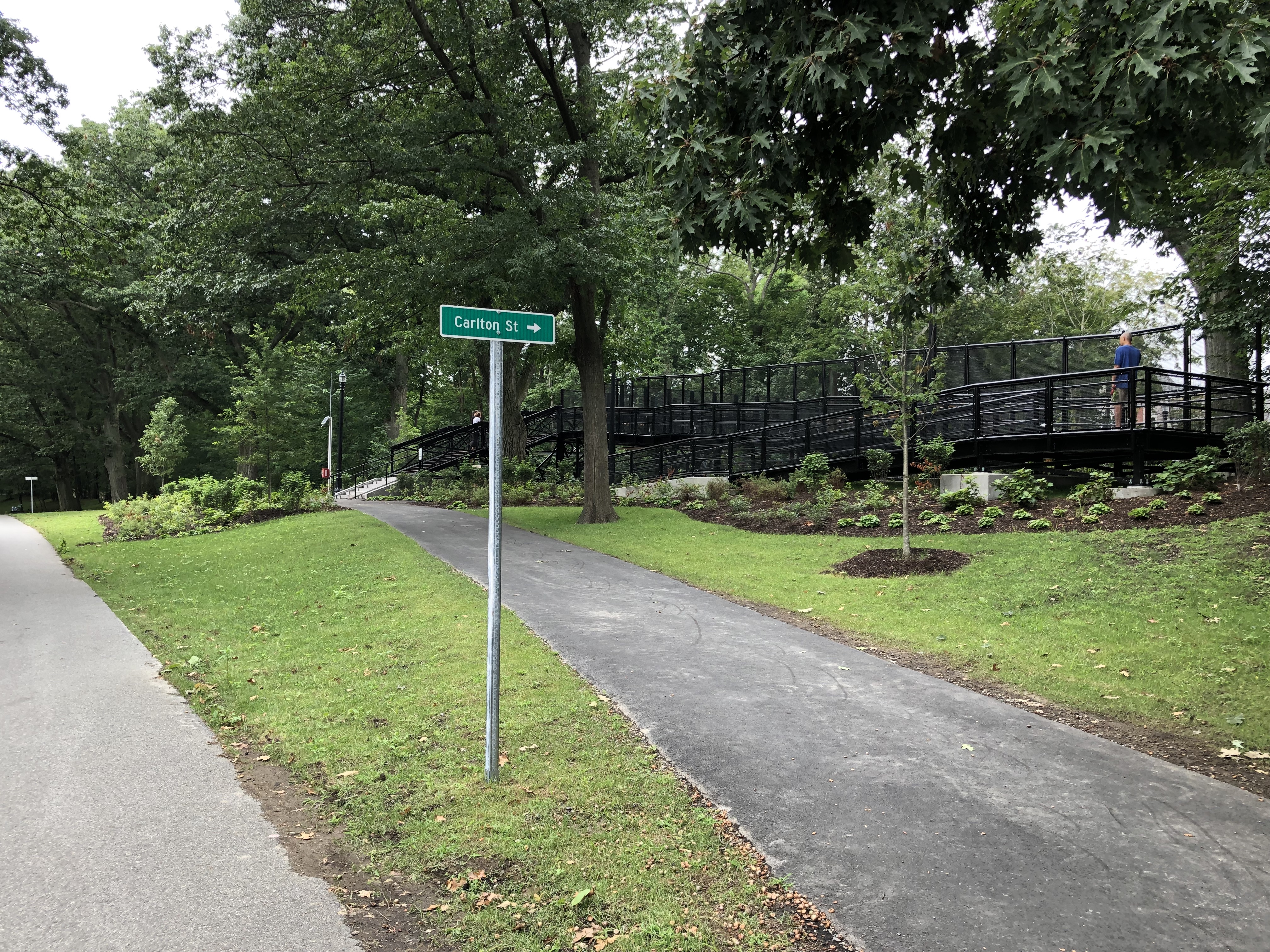 Two paved paths diverge in a green woodland. A green street sign pointing right indicates that the right path leads to Carlton St. At the top of that path, a stairway and a wheelchair ramp made of black metal lead over the Green Line tracks.