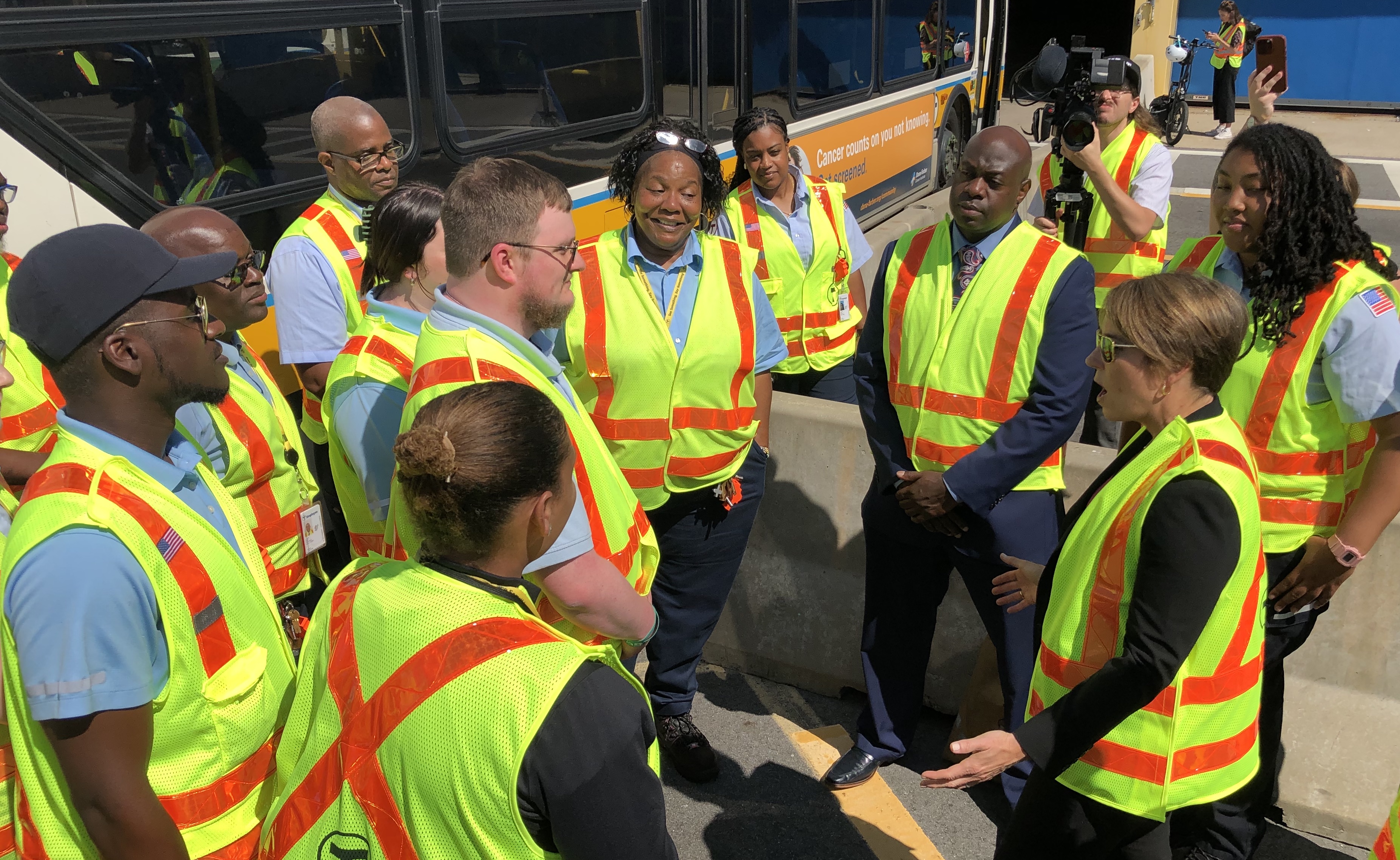 A group of people wearing fluorescent yellow safety vests chats in a circle next to a parked MBTA bus.