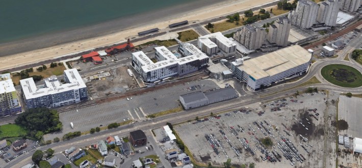 An aerial view of an oceanfront neighborhood dominated by large surface parking lots and a handful of mid-rise apartment buildings lining the beach in the upper left corner of the image.