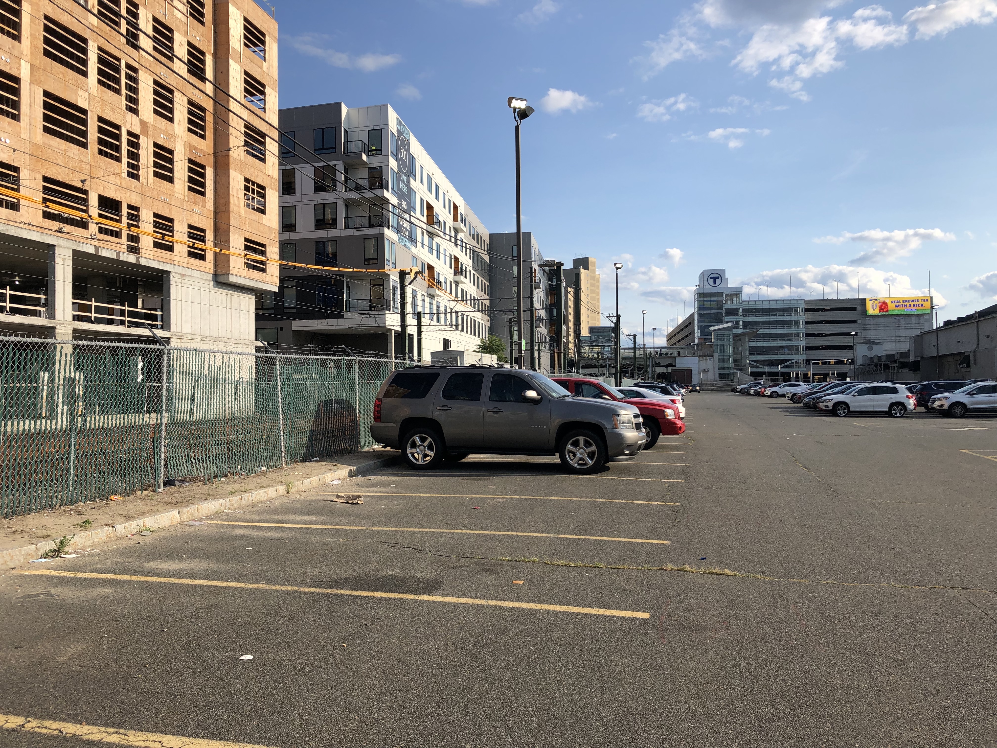 A half-empty parking lot next to several mid-rise apartment buildings, one of which (on the left edge of the photo) is still under construction. In the distance is a large parking garage with the MBTA logo on top. Some railroad tracks – the MBTA Blue Line – are visible through a chain-link fence at left, between the apartments and the parking lot.
