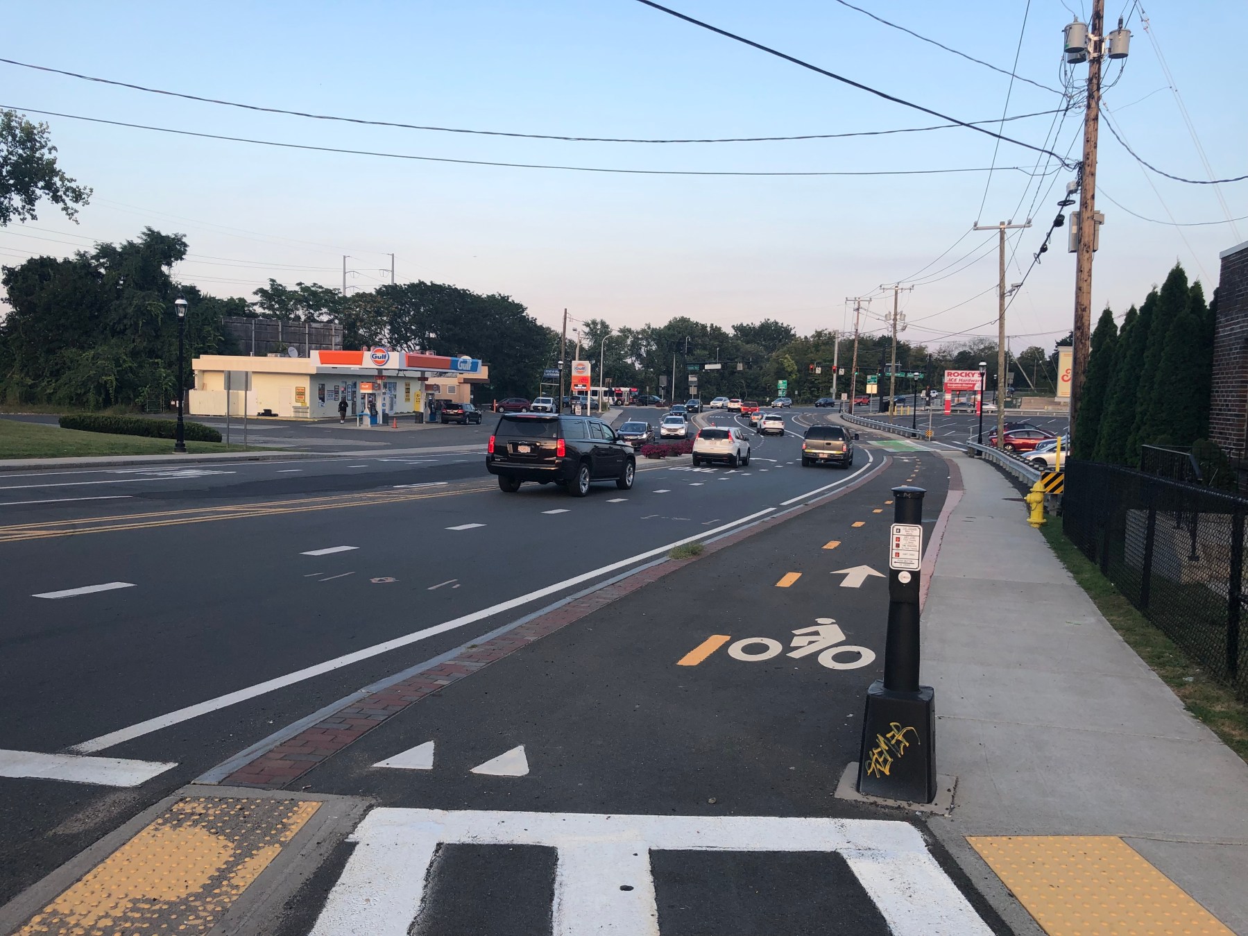 A paved bike path runs alongside a wide multi-lane road (at left) and a sidewalk (at right) with a striped crosswalk in the foreground. To the left of the roadway is a gas station and to the right is a parking lot.