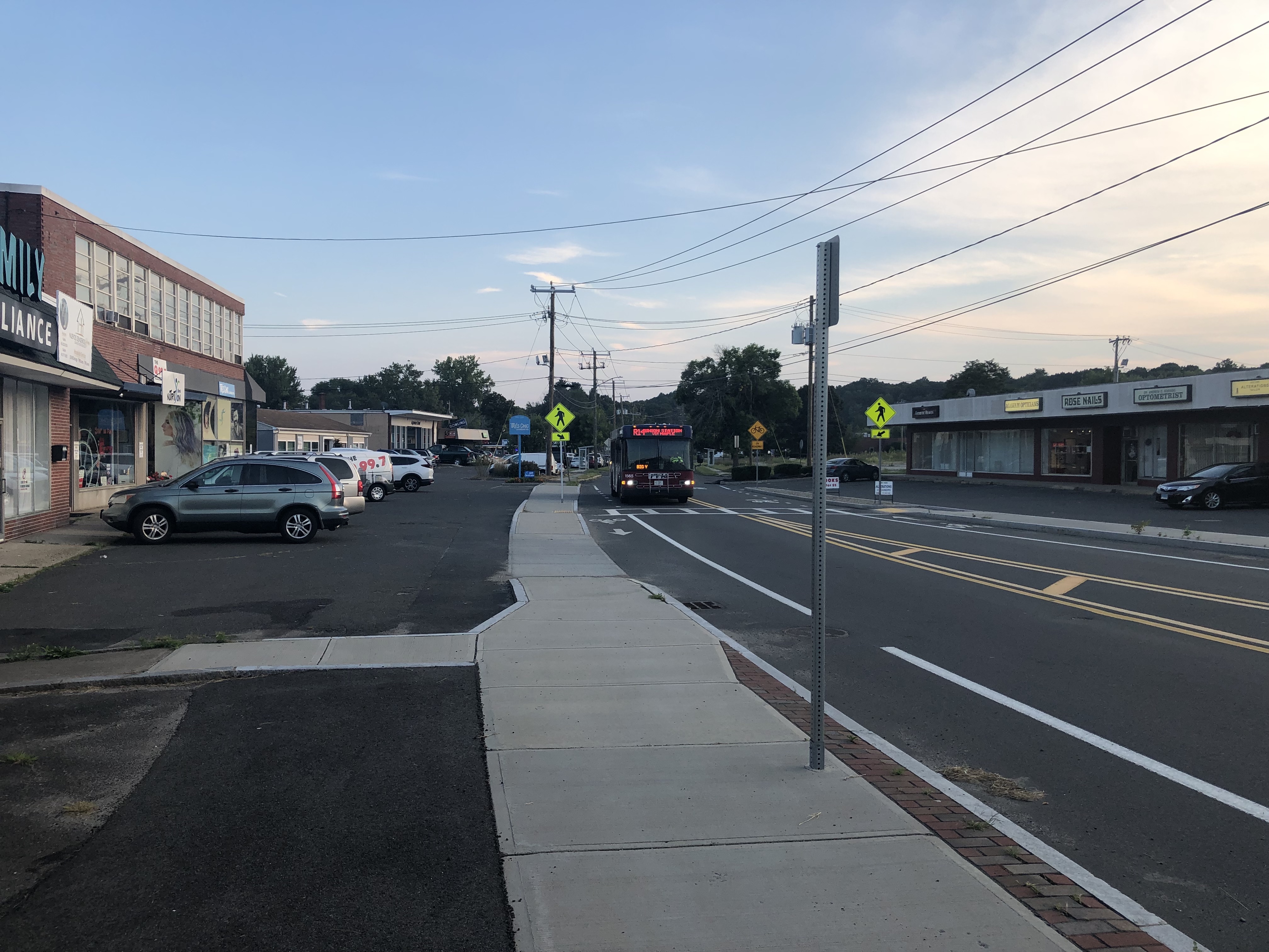 A PVTA bus approaches along a two-way street lined with strip malls and parking lots. The street includes new sidewalks and painted bike lanes and the strip malls on either side are full of small businesses.