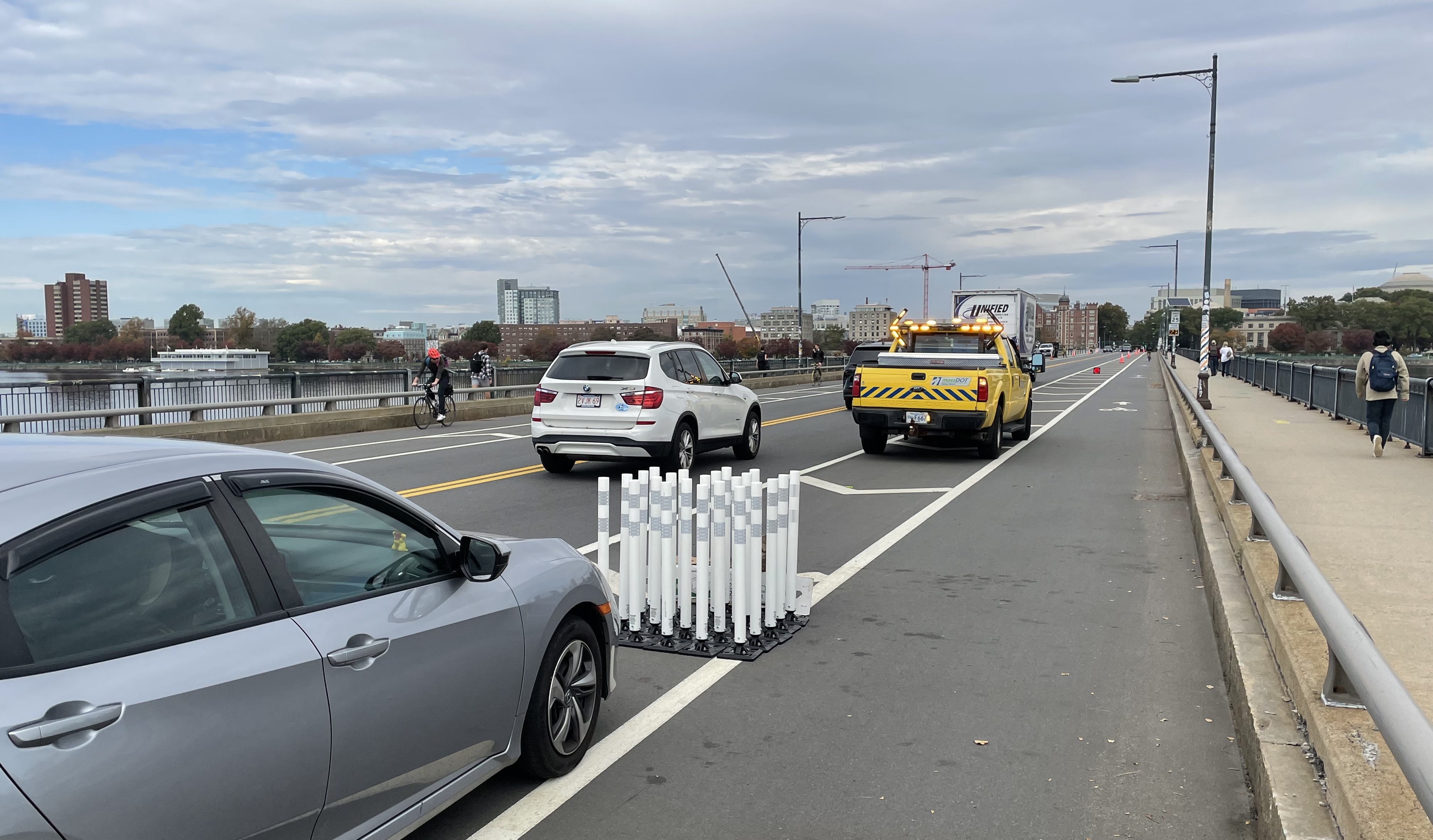 A photograph of a bridge with a bike lane and wide sidewalk at right, and two MassDOT vehicles in the buffer zone between the car lane and the bike lane. A cluster of white flexible-post bollards is bundled between the two vehicles. In the distance, at the end of the bridge, is the Cambridge skyline.