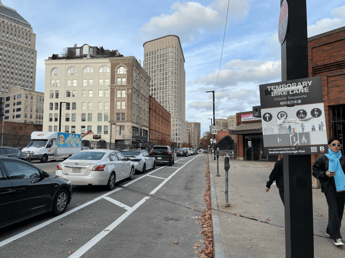 A protected bike lane crosses a viaduct between a sidewalk and a row of parked cars, with several tall buildings visible along the other side of the street in the distance. On the right is an old sign that says "Temporary Bike Lane - Ciclovia Temporaria – for more information visit boston.gov/orange-line". A sign further in the distance marks this street as Massachusetts Route 28.