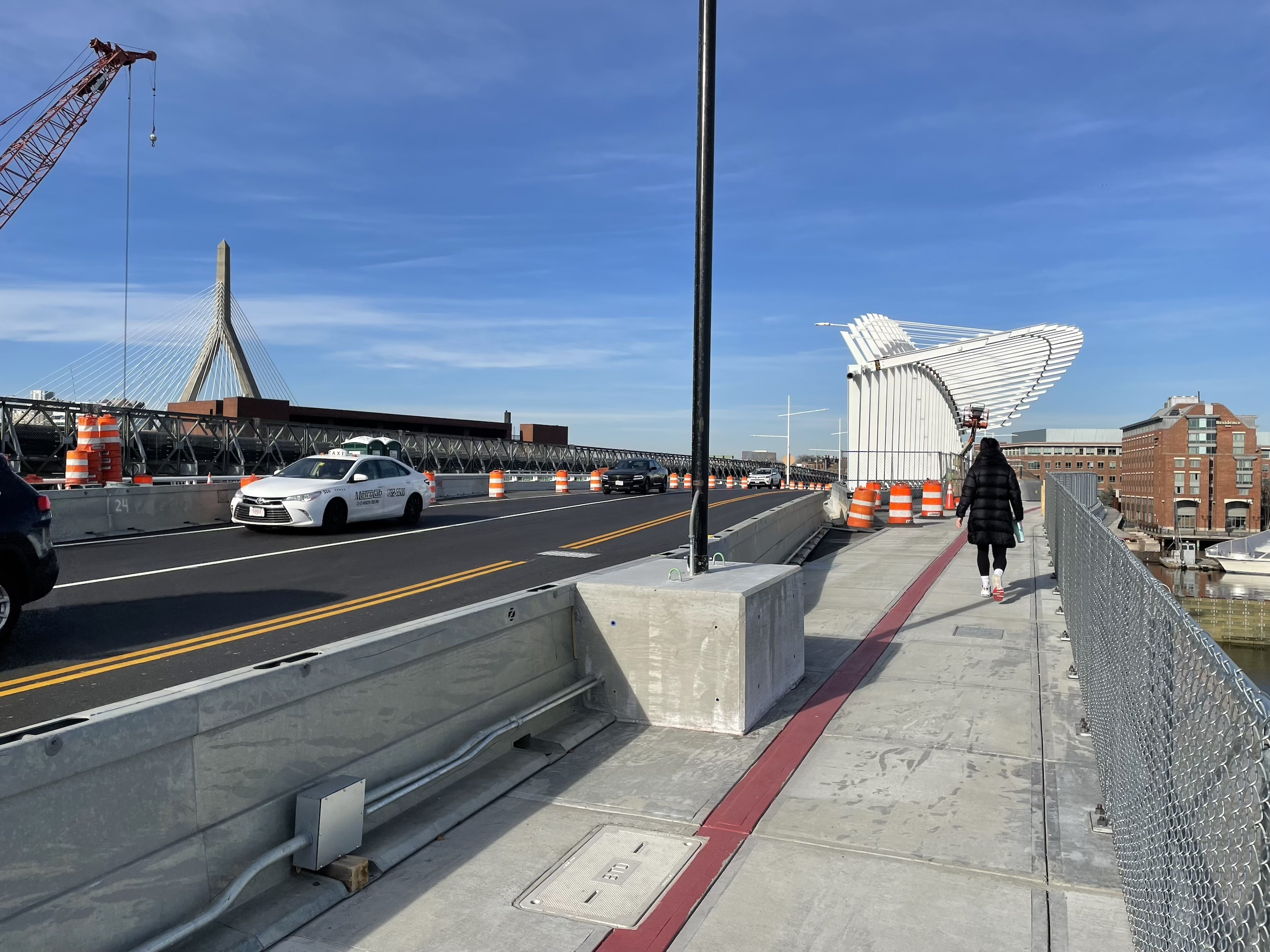 A pedestrian walks on a sidewalk next to some temporary crash barriers on a new bridge. To the left several cars drive on fresh asphalt, and on the left edge a crane is visible among some orange construction barrels.
