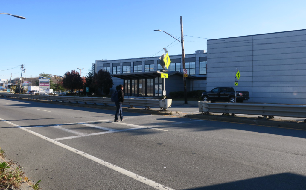 A man in a sweatshirt crosses a four-lane highway with a crash barrier guardrail in the center. On the opposite side of the street is a gray warehouse building.