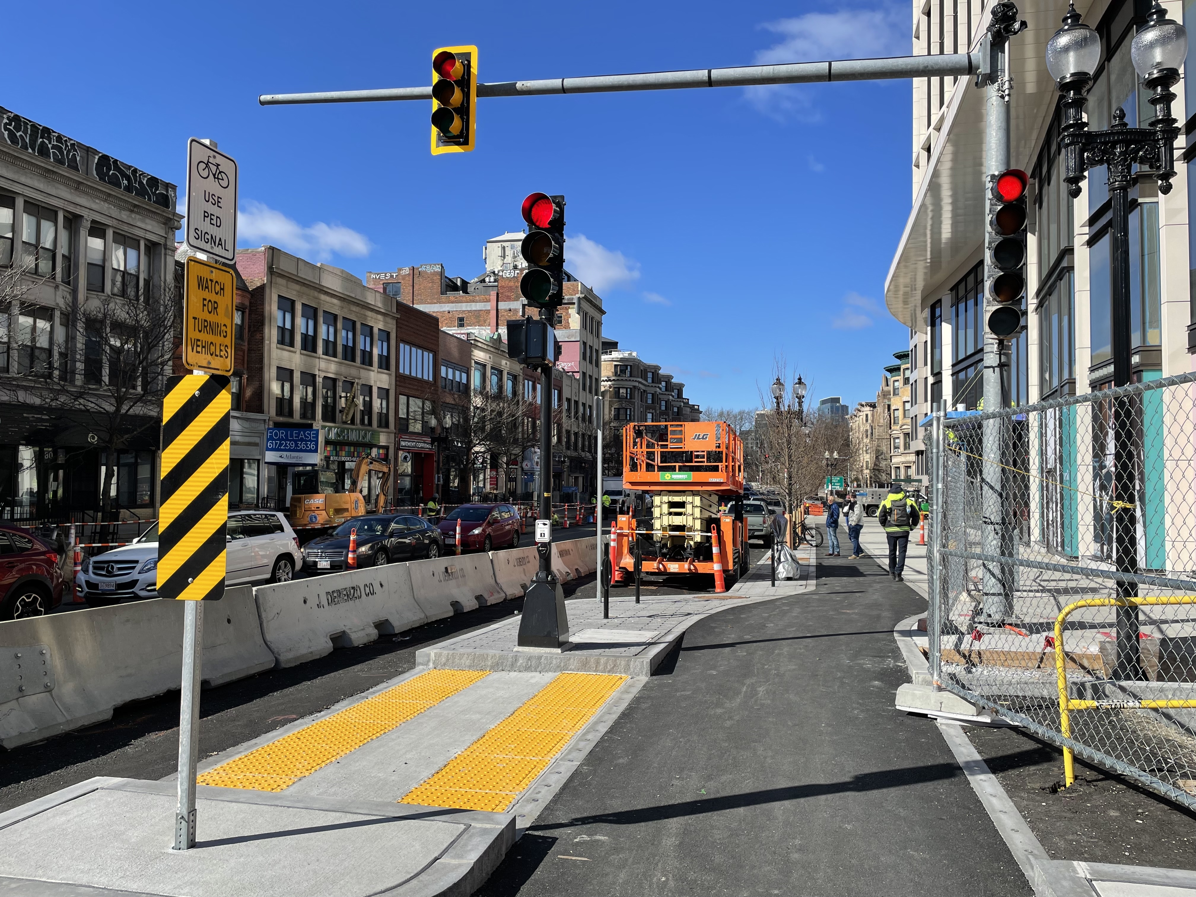 A new sidewalk, new traffic lights, and a protected bike lane run along a construction site. On the opposite side of the street are several older multi-story brick buildings.