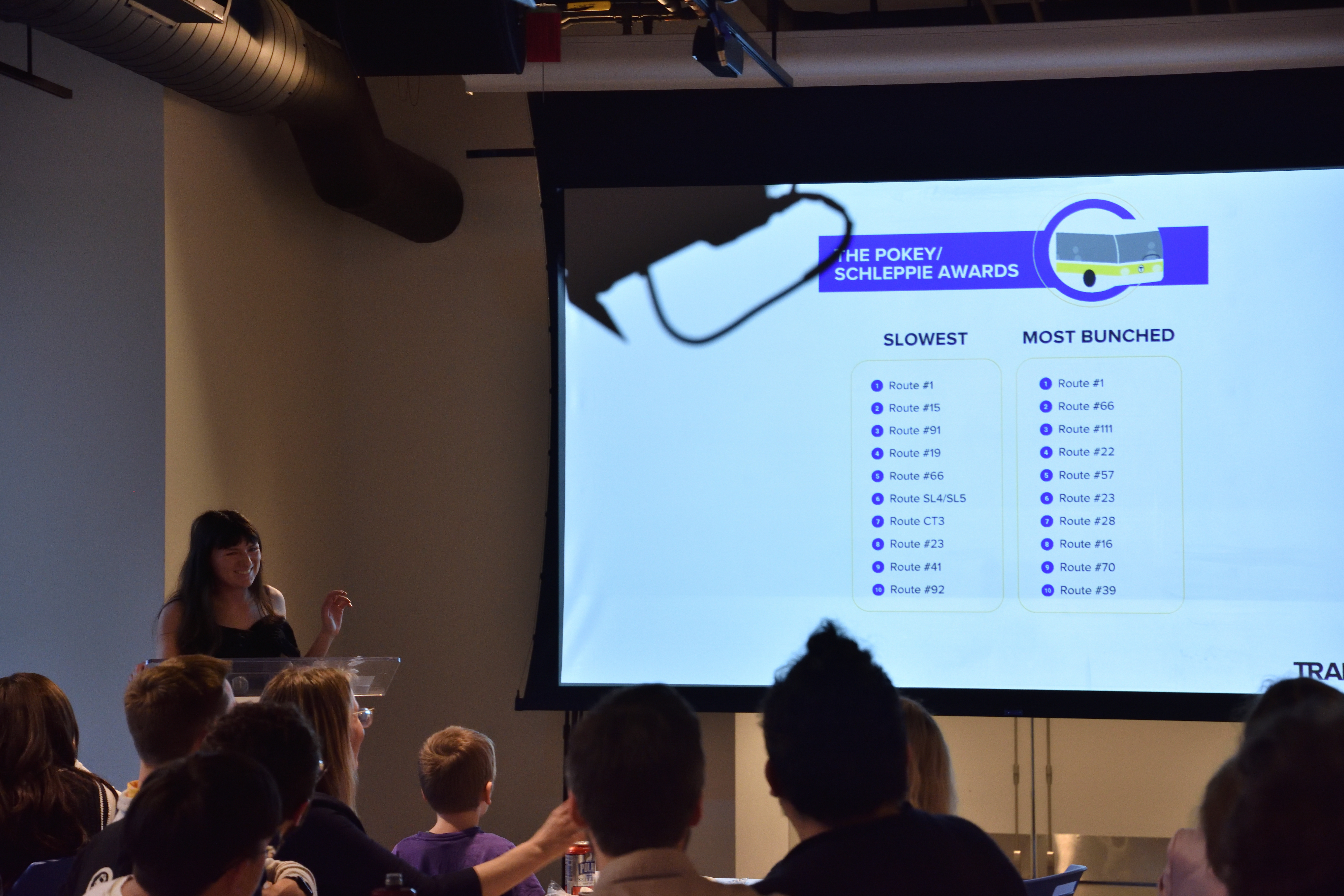A woman in a black dress standing at a podium presents a list of MBTA bus routes projected on a screen in front of a crowded room of audience members. 