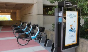 A bikesharing station kiosk with four parked bikes in a sheltered passageway of a concrete building. The kiosk has a sign that says "Holyoke Community College" and an ad that says "ride an electric-assist bike"