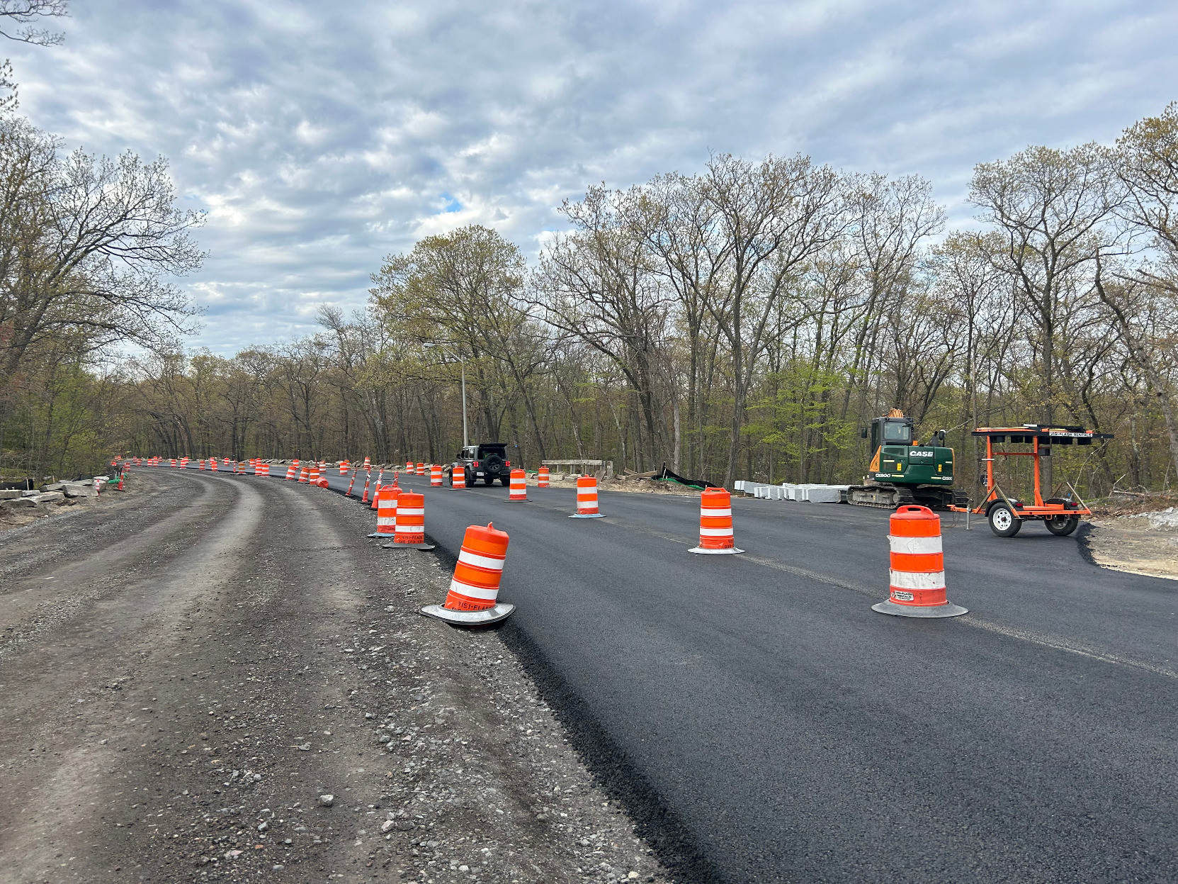 A photo of a street construction site running through woods that are just beginning to leaf out with spring leaves. On the left side of the photo is a roughly 20-foot-wide expanse of dirt and gravel. To the right is a freshly-paved two-lane road, lined with orange construction barrels. On the far side of the pavement is an excavator and an orange construction trailer.