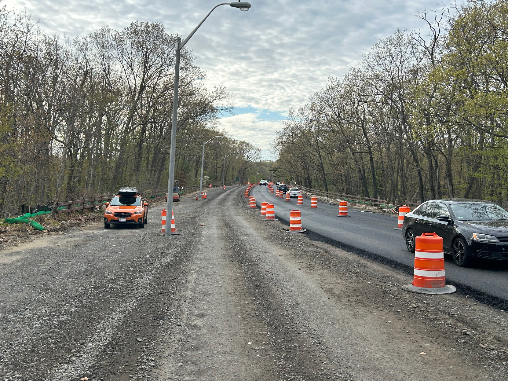 A dirt track runs alongside a freshly-paved two-lane road on the right side of the photo. The edge of the pavement is lined with orange construction barrels. The road is surrounded by trees just beginning to grow their leaves.