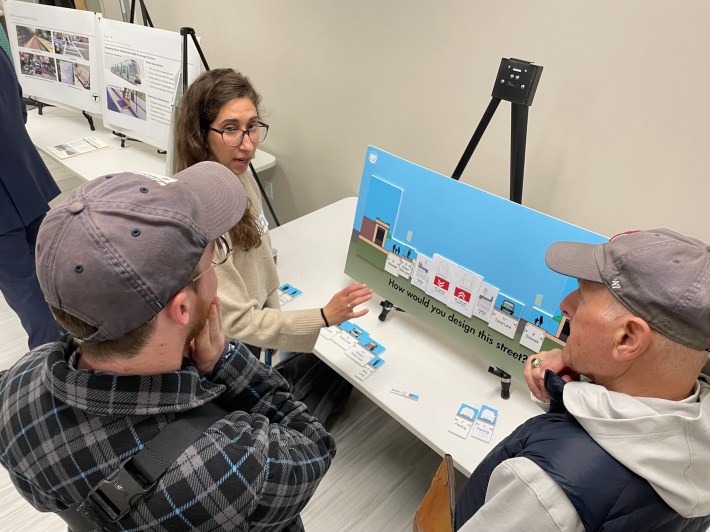 Two men in baseball hats and a younger woman with glasses cluster around a magnetic board that represents a cross-section of a street. Magnets stuck to the board allow them to rearrange elements of the street. The board shows two red transit lanes in the middle of the street, two boarding platforms on either side, a vehicle lane in each direction outside of the platforms, and sidewalks on the edges.