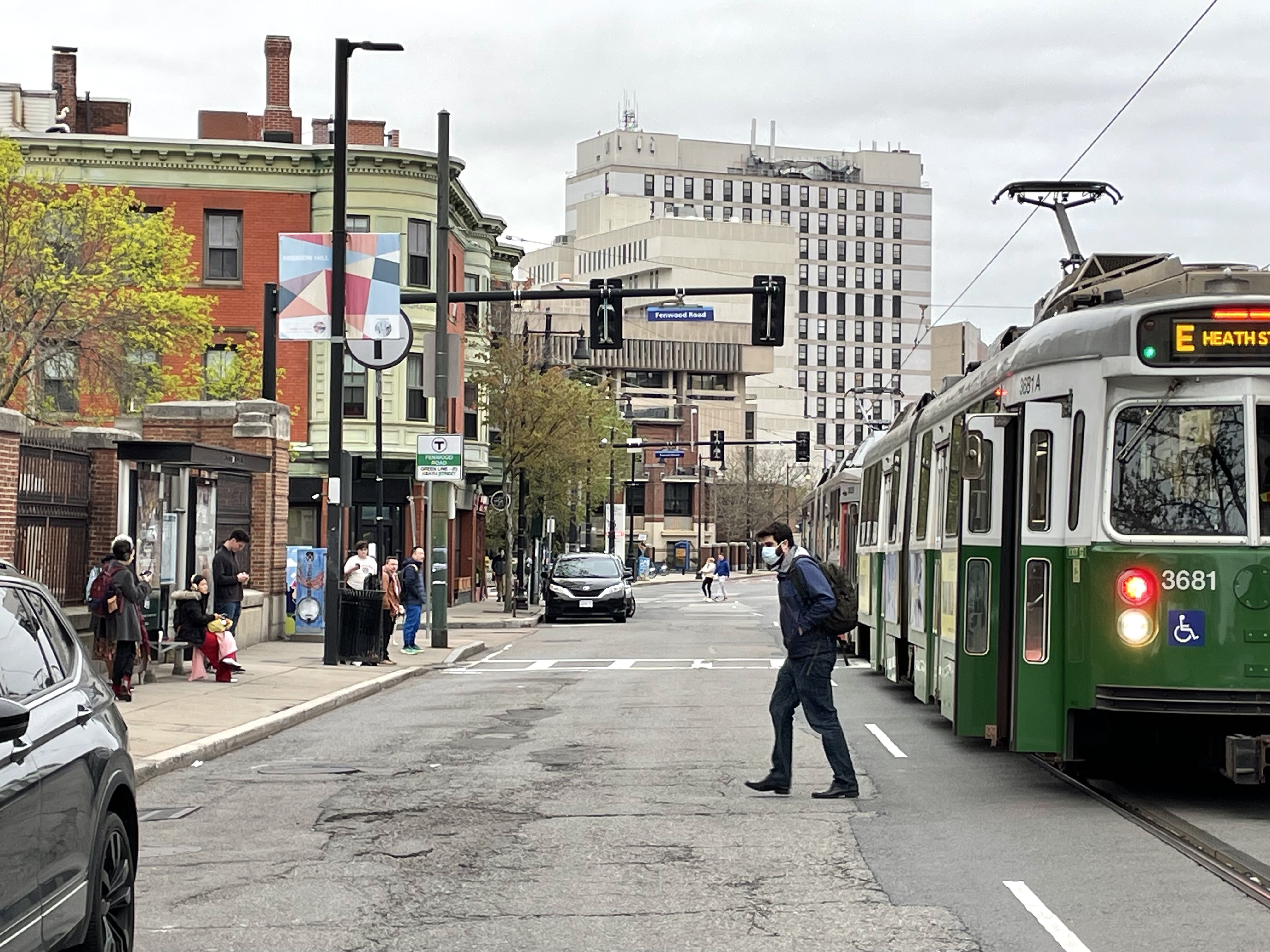 A green line train stops in the middle of Huntington Ave. while a passenger disembarks in the middle of the roadway while other riders wait under a bus shelter on the sidewalk at left.