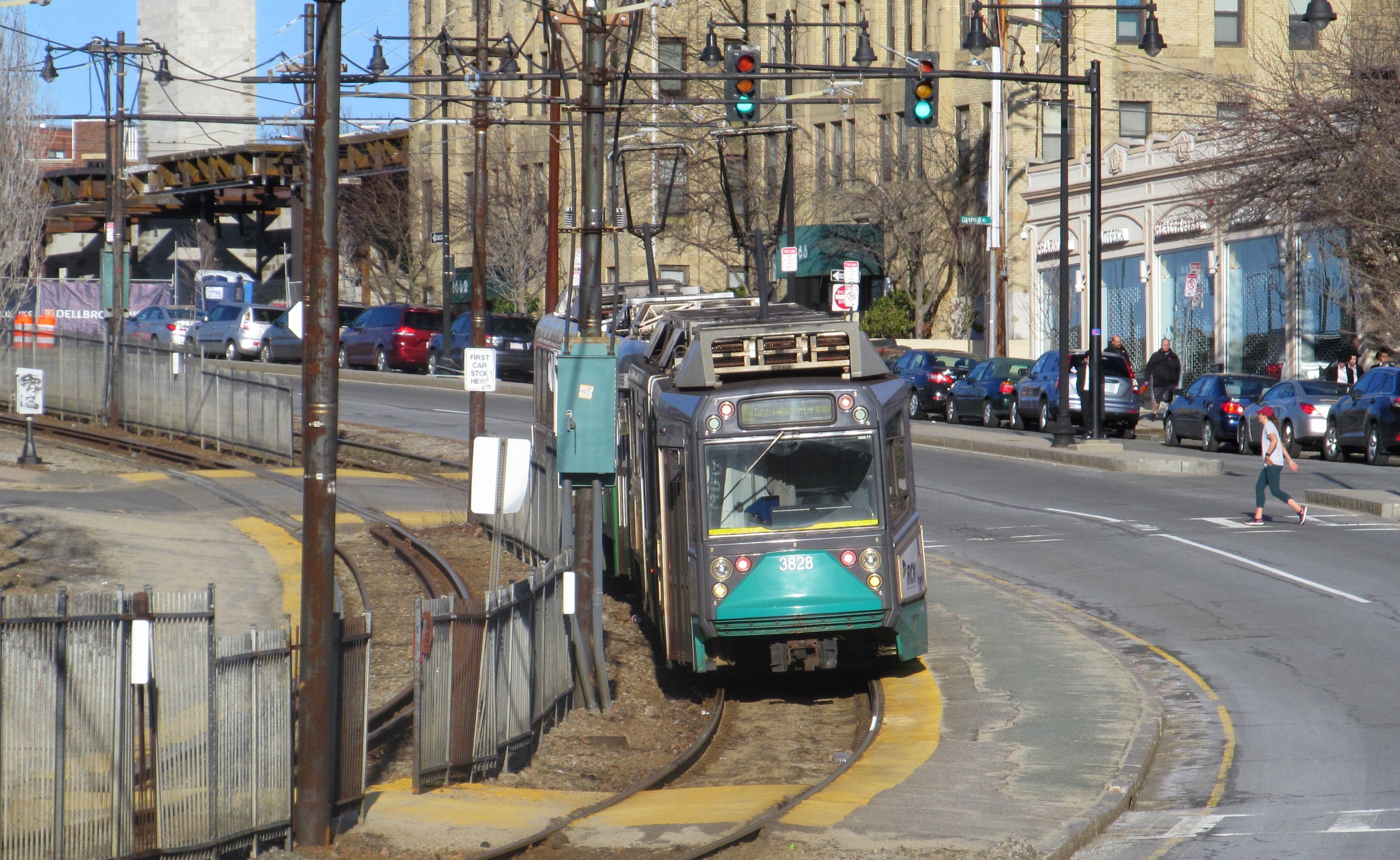 A green line train stops in the median of a multi-lane roadway as a pedestrian crosses in a crosswalk towards it.