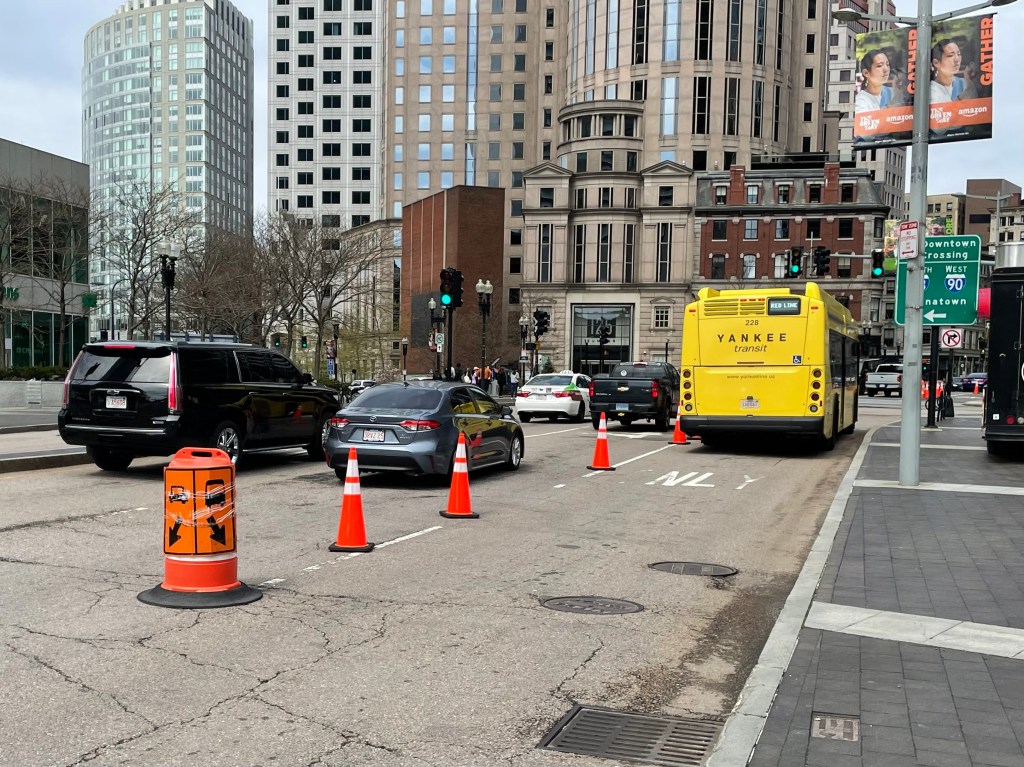 A yellow shuttle bus at right drives in a lane separated from other traffic by a row of orange construction cones. A orange barrel in the left foreground shows a sign with a bus icon pointing to the right and a car icon pointing left. High rise office buildings are in the background