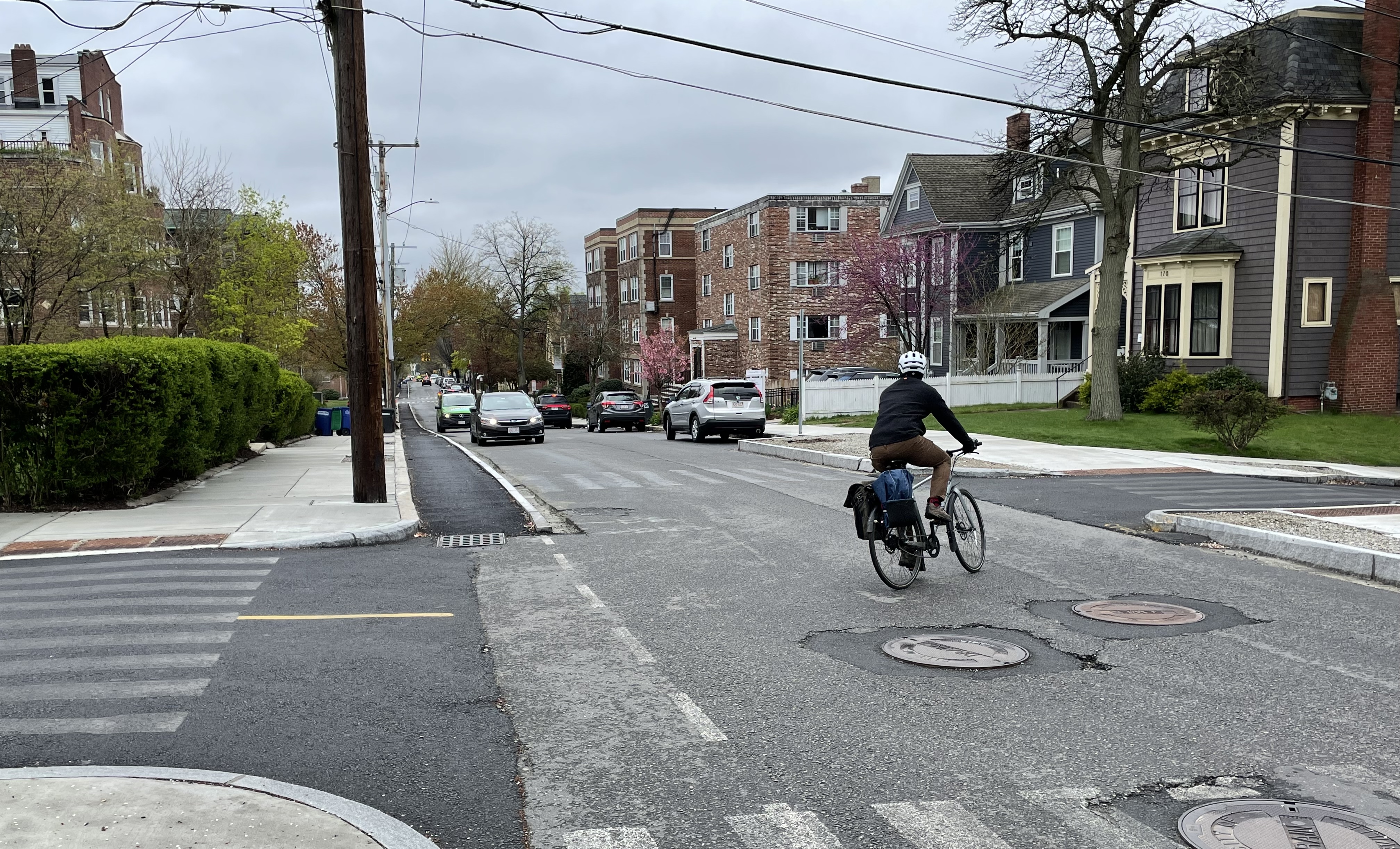 A bicycle rider turns onto a street between two fresh patches of pavement. To their left, a partially-paved, curb-protected bike lane stretches downhill into the distance along the street's left curb, as two cars drive up the hill in the middle distance, approaching the camera. The street is lined with blossoming trees and multi-story apartment buildings, with several larger 4- and 5- story brick buildings in the distance.