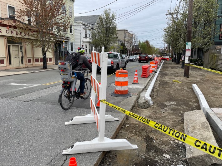 A man riding a bike with a child's seat on the back pedals past a construction zone marked with cones. To his right, fenced off with barriers, is a gravel lane delineated with granite curbs. A line of cars stopped in traffic waits in the distance. 
