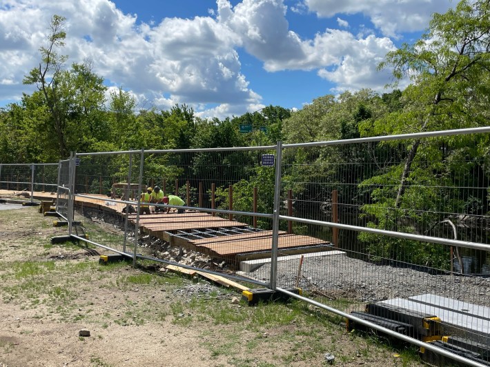 Workers behind a metal fence install planks on a new boardwalk along a tree-lined river. 