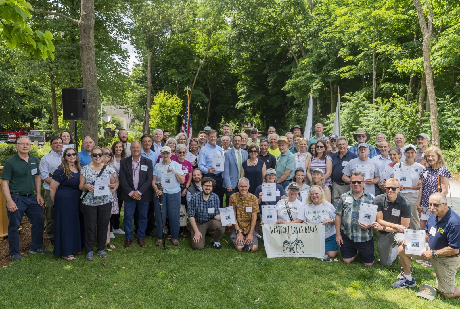 A crowd of people on a grassy lawn surrounded by trees smiles for the camera while holding up certificates.