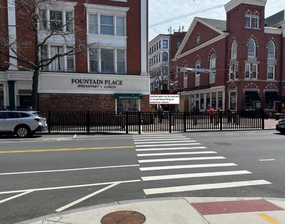 A rendering of a city street with a black metal fence surrounding the sidewalk on the far side of the street. In the background is a brick multi-story building