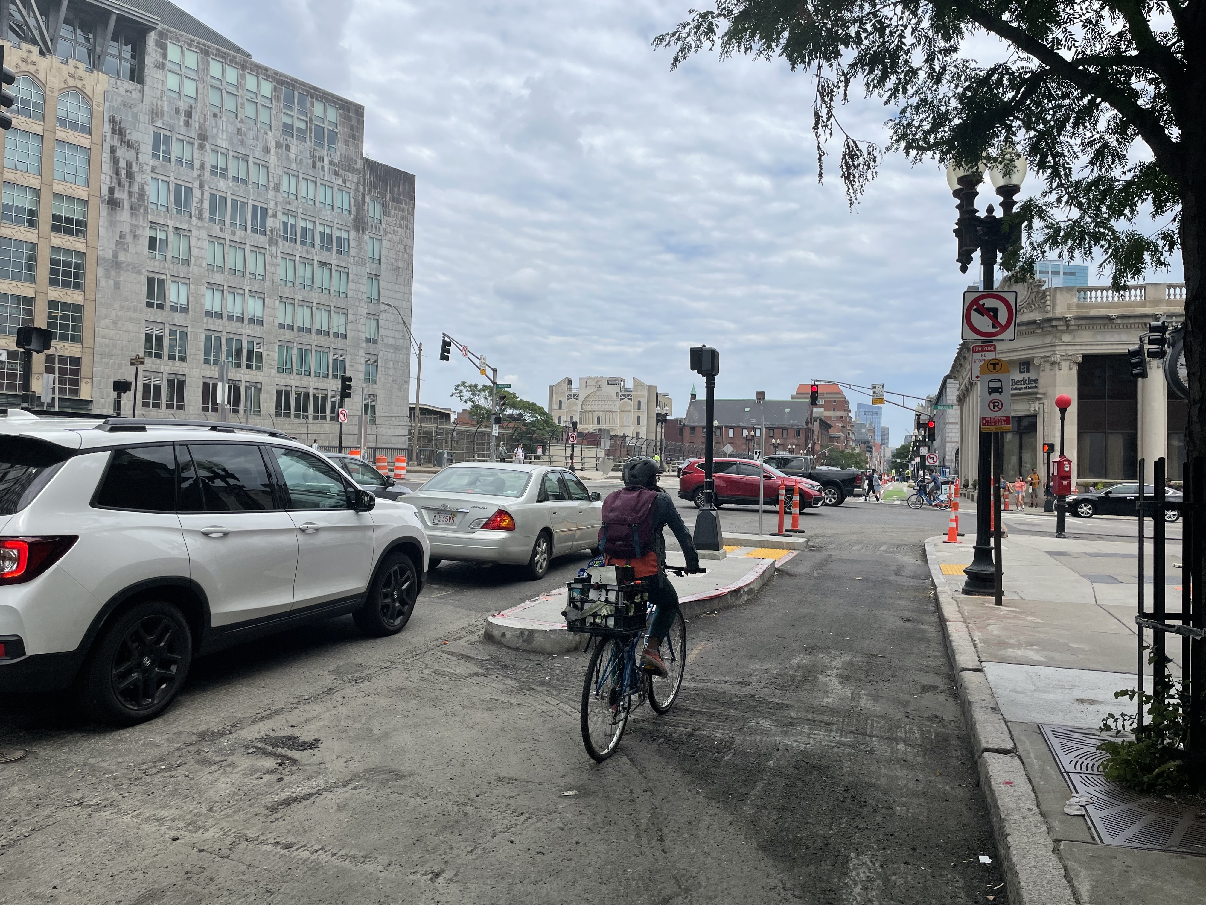 A person on a bike approaches an intersection in a bike lane that's separated from the adjacent car lane by a granite curbed island with a walk signal. The street is surrounded by high-rise buildings and there are several orange construction cones in the intersection.