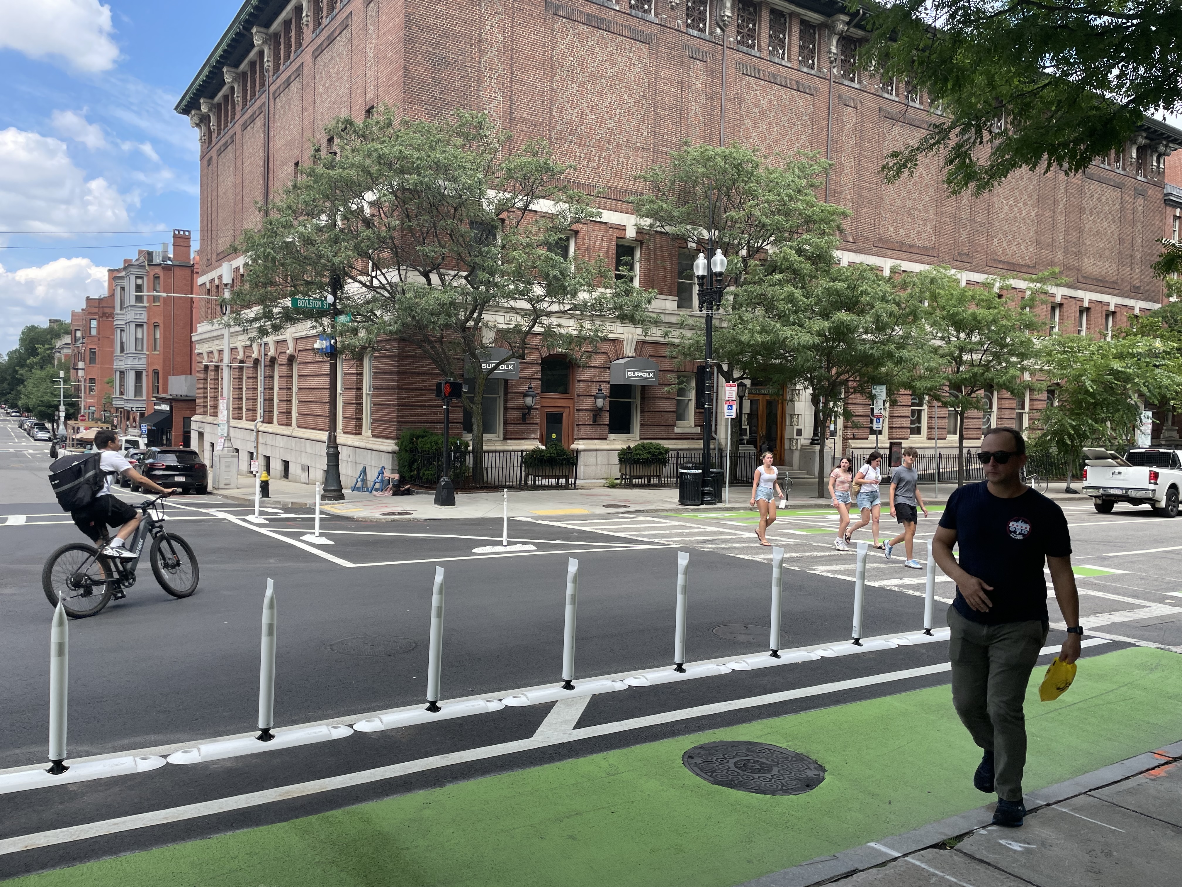 A person on a bike and five pedestrians cross a street lined with flexible-post bollards that separate a green bike lane in the foreground from motor vehicle lanes in the middle of the street. In the background is a large brick building.
