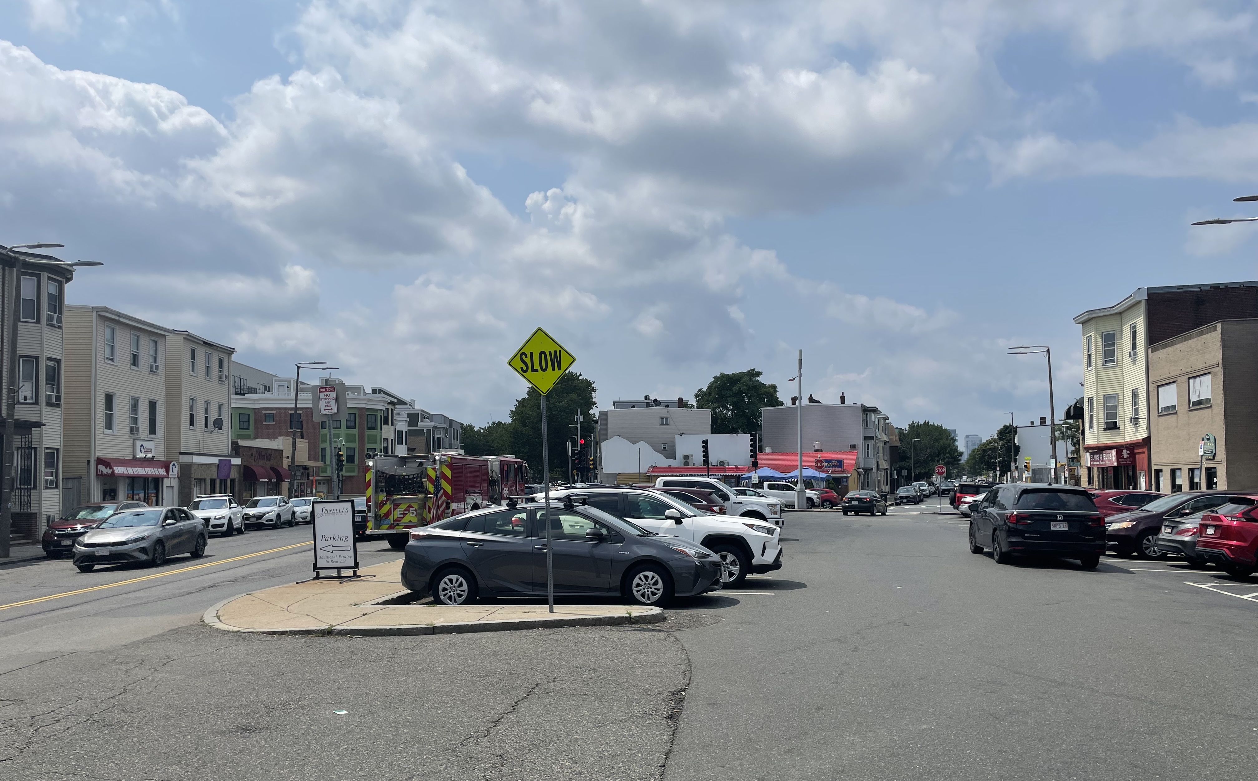 A large parking lot next to a two-lane city street, surrounded by 3- and 4-story buildings. In the foreground is a diamond-shaped "SLOW" sign