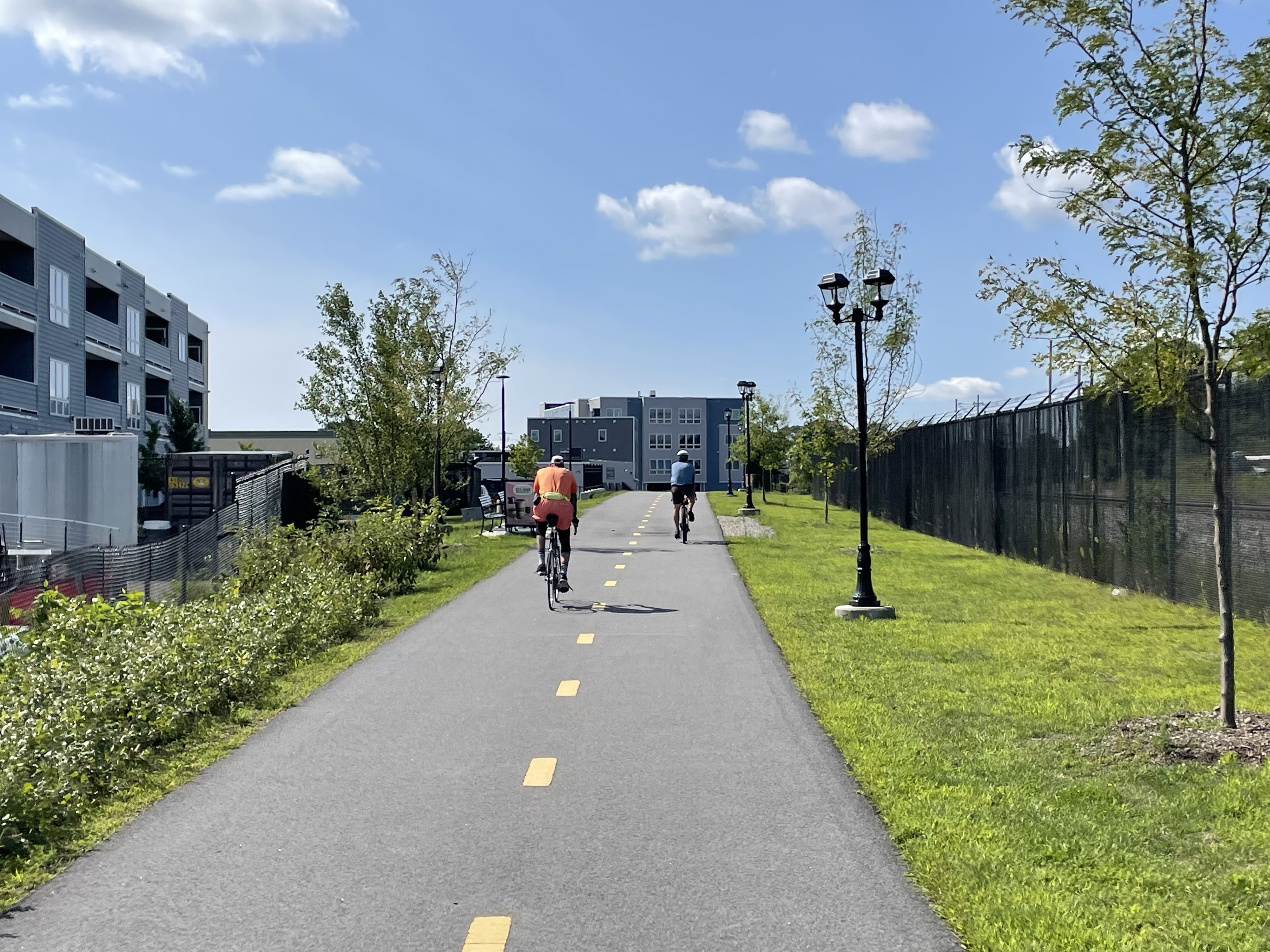 Two people on bikes ride along a straight bike path between a fence and some decorative light posts on the right and some mid-rise new apartment buildings to the left.