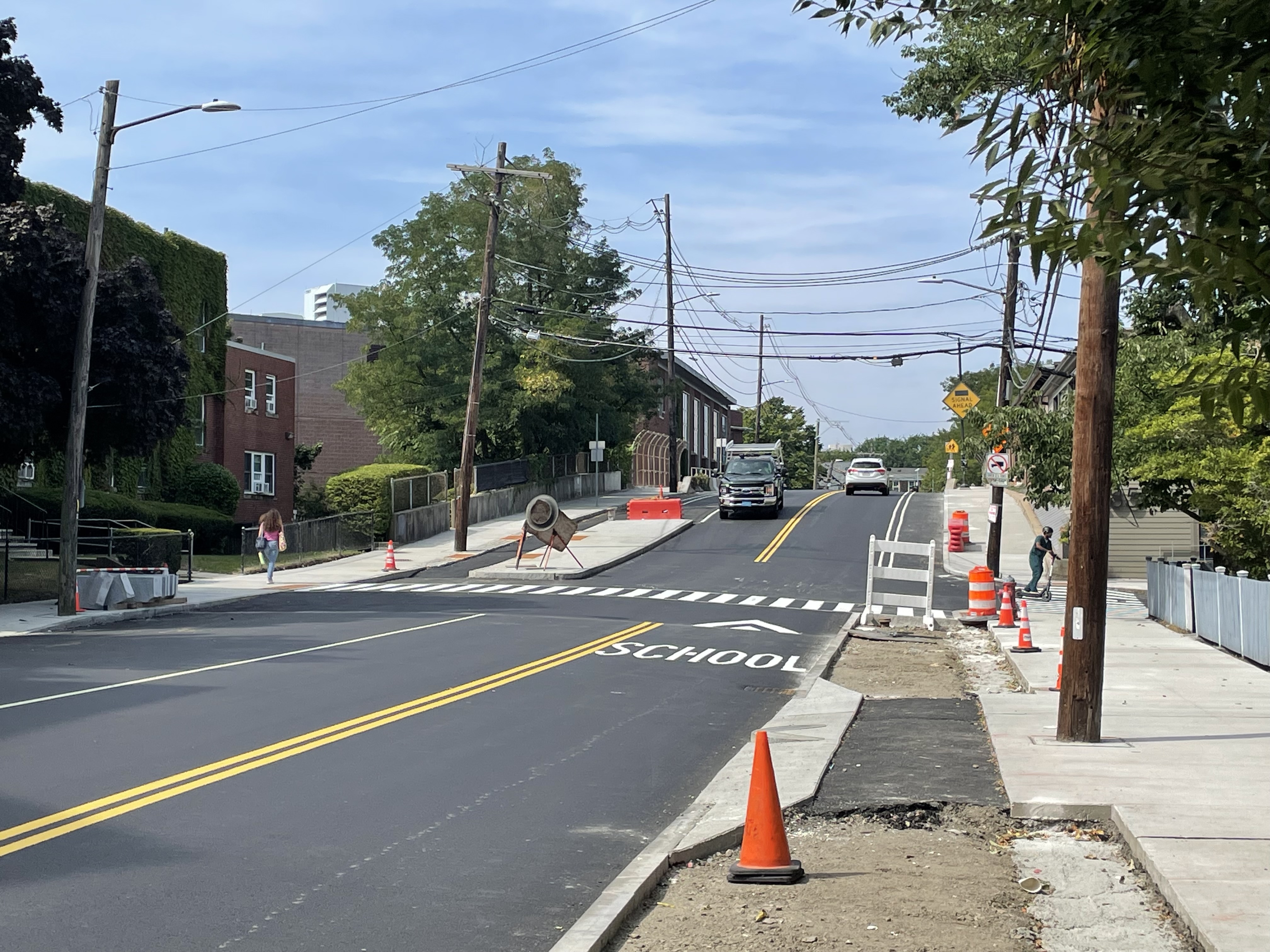 A newly paved street with orange construction barrels and cones along the curb in the foregound, where the sidewalk and bike lane are still under construction.