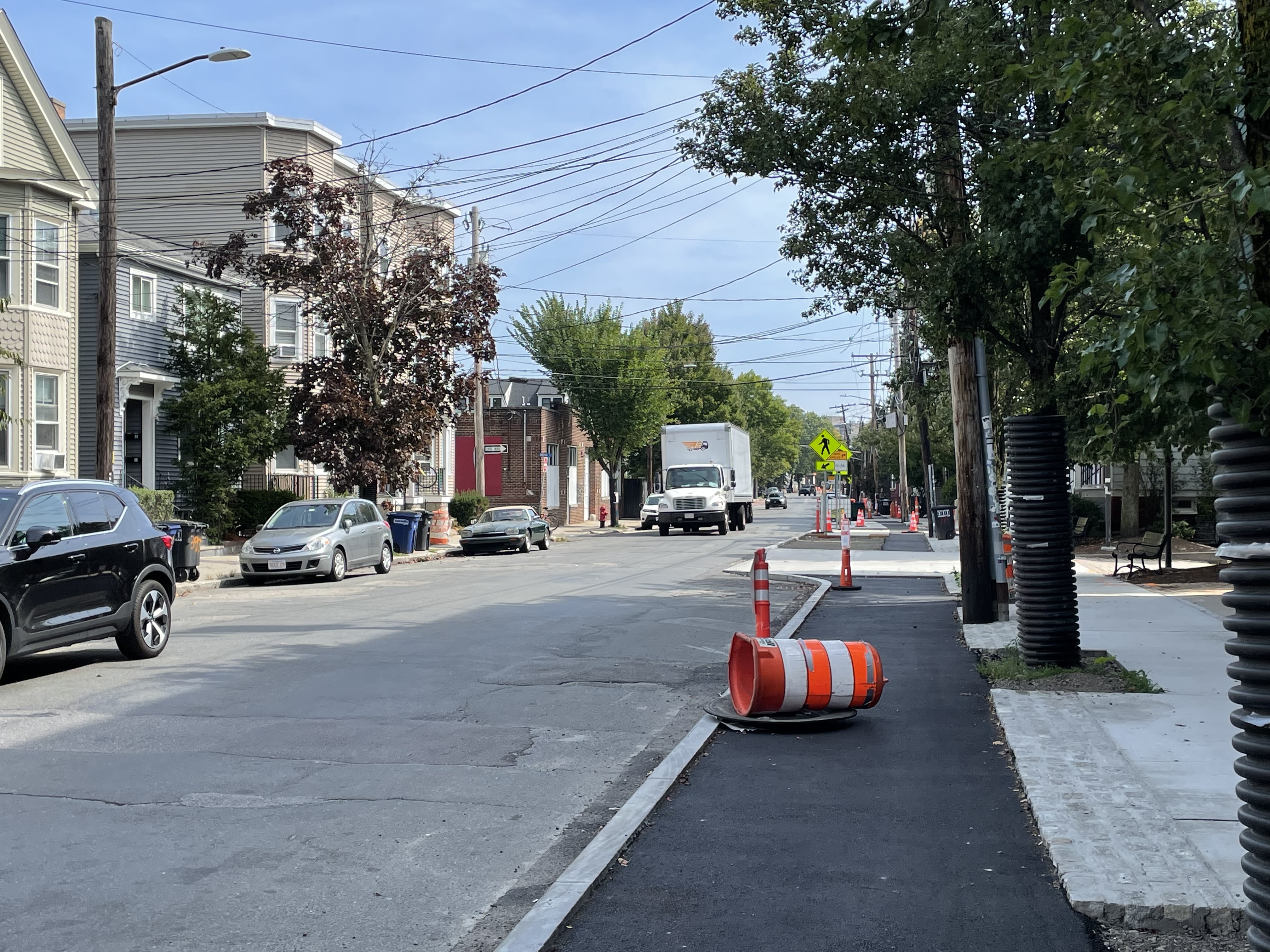 A new sidewalk-level bike lane under construction adjacent to a two-lane city street with a few cars parked along the opposite curb. Cones and construction barrels litter the bike lane which is still awaiting a final layer of pavement.